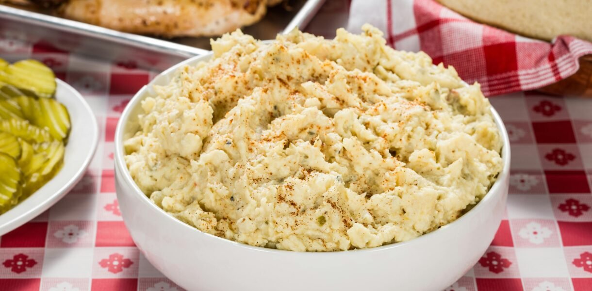 Creamy potato salad in a white bowl with BBQ and pickles on a checkered tablecloth.