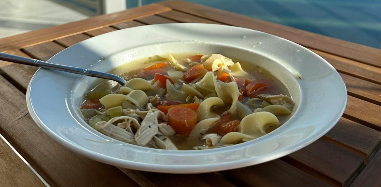 A bowl of chicken noodle soup on a balcony table overlooking a serene waterfront cityscape.
