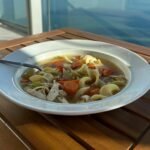 A bowl of chicken noodle soup on a balcony table overlooking a serene waterfront cityscape.