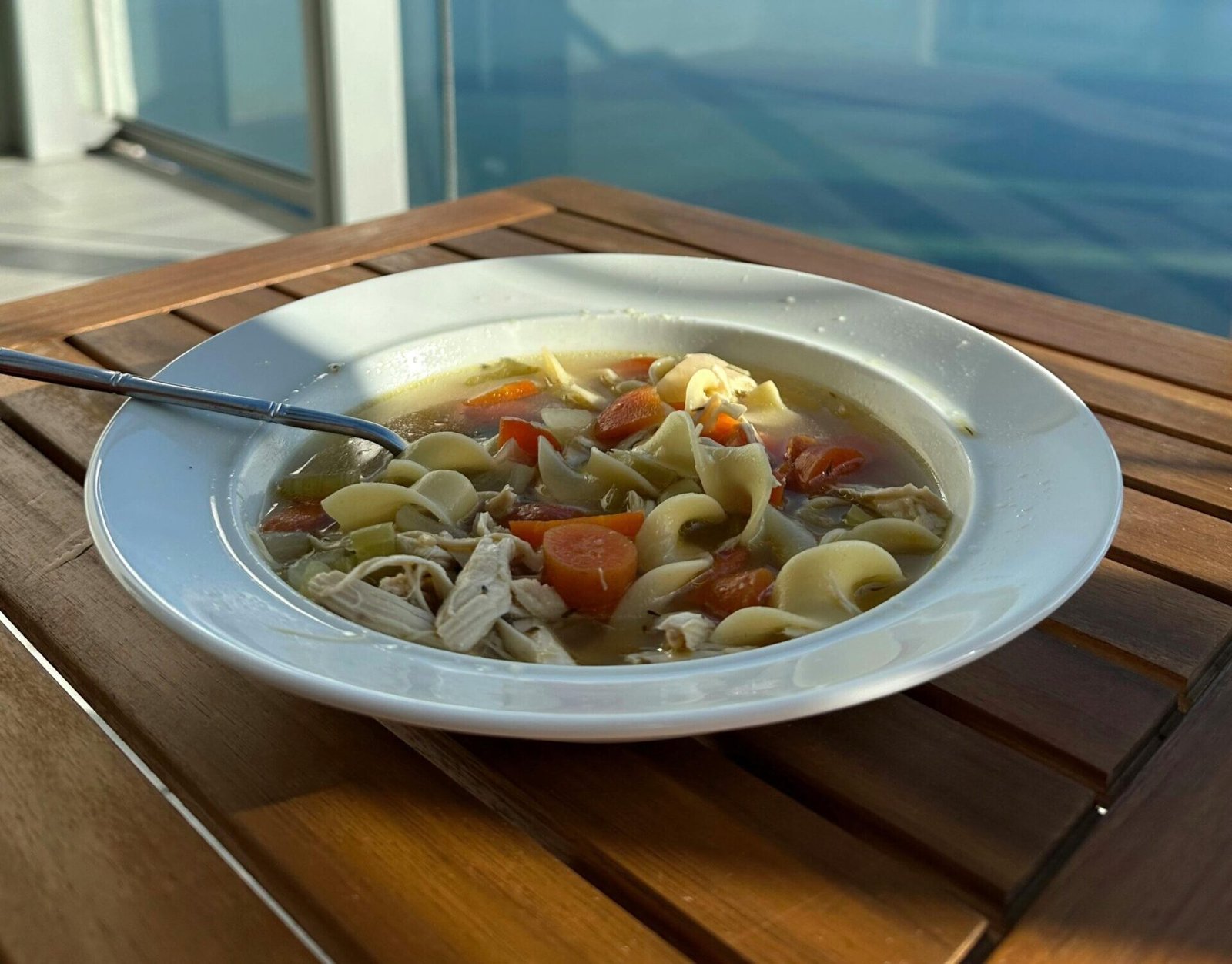 A bowl of chicken noodle soup on a balcony table overlooking a serene waterfront cityscape.