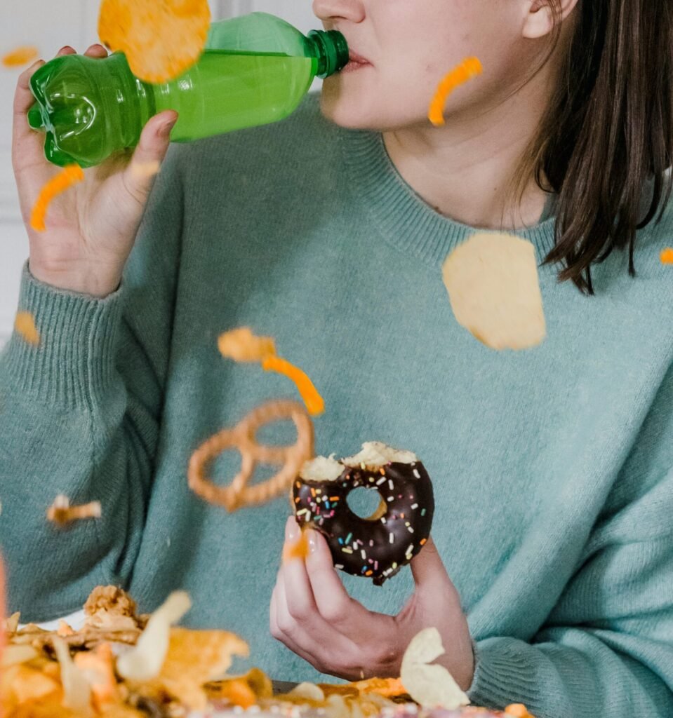 Delighted female with closed eyes showered by chips and crisps drinking fresh cold soda and eating yummy donut near heap of yellow snacks